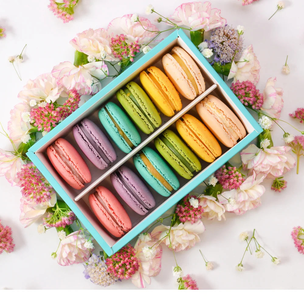 Box of colorful Birthday Macarons arranged in rows, placed on a surface adorned with delicate pink and white flowers, perfect for a special occasion or gift.