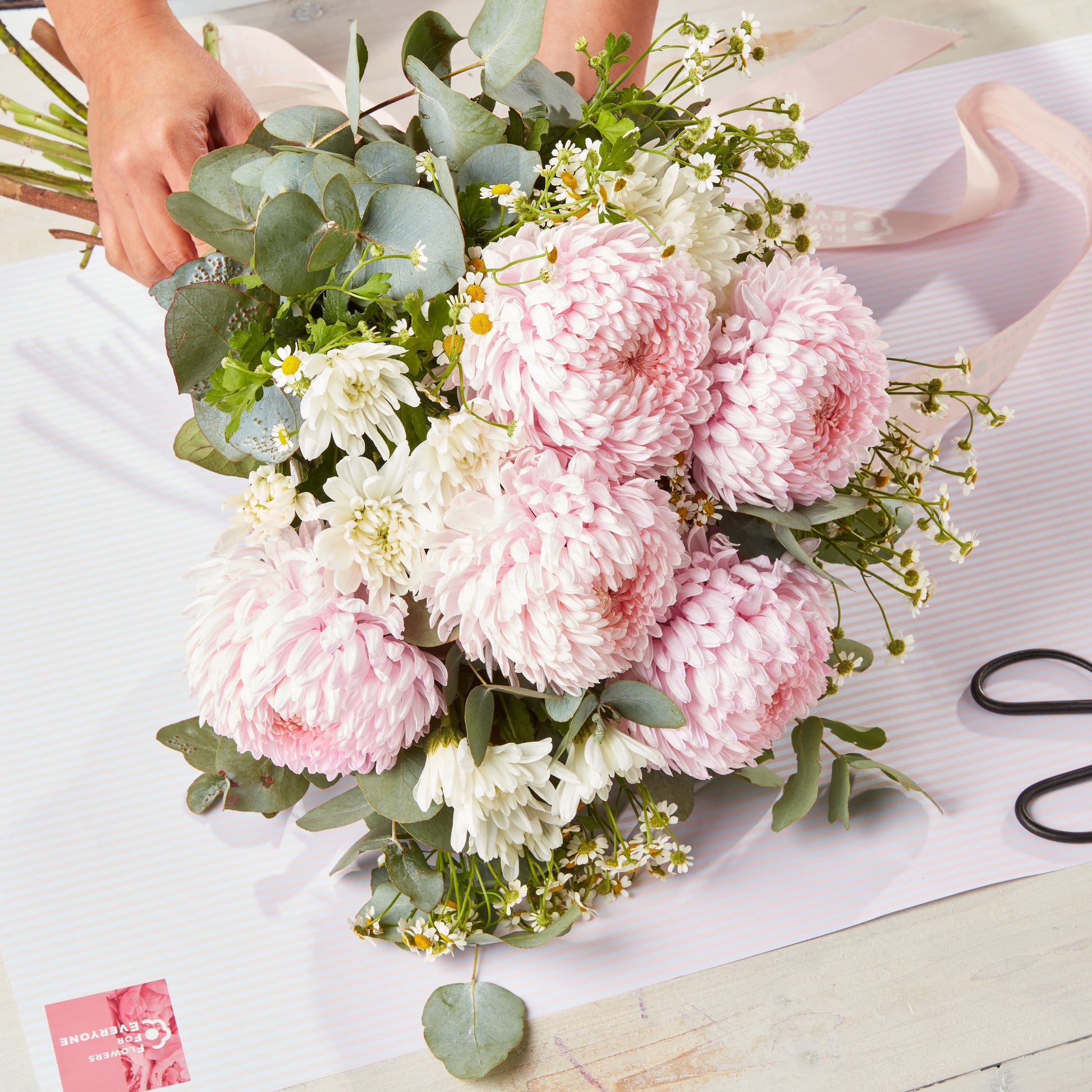 Hand holding Pretty in Blush Bouquet featuring soft pink chrysanthemums, white daisies, eucalyptus leaves, and small white filler flowers on a white striped surface.