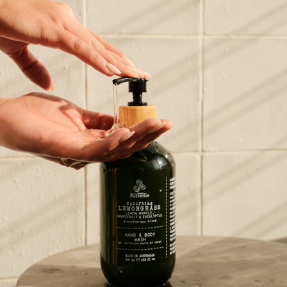 The Wellness Ritual hand and body wash bottle with a pump being used under warm sunlight in a tiled bathroom