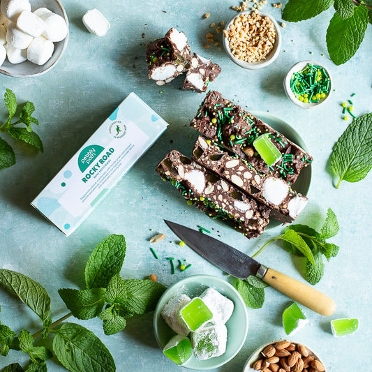 A styled flat-lay of a chocolate bark bar product with mint, almonds, marshmallows, and a knife on a light blue surface. The box reads 'Minty Mint Chocolate Bark' with a green logo.
